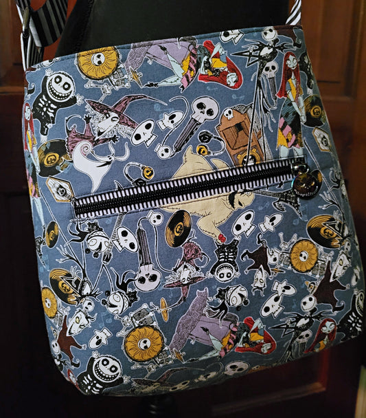 A fabric tote bag featuring a dense cartoon print of Jack Skellington, skeletons, and other spooky characters on a muted blue-gray background, with a black-and-white striped zipper across the front.